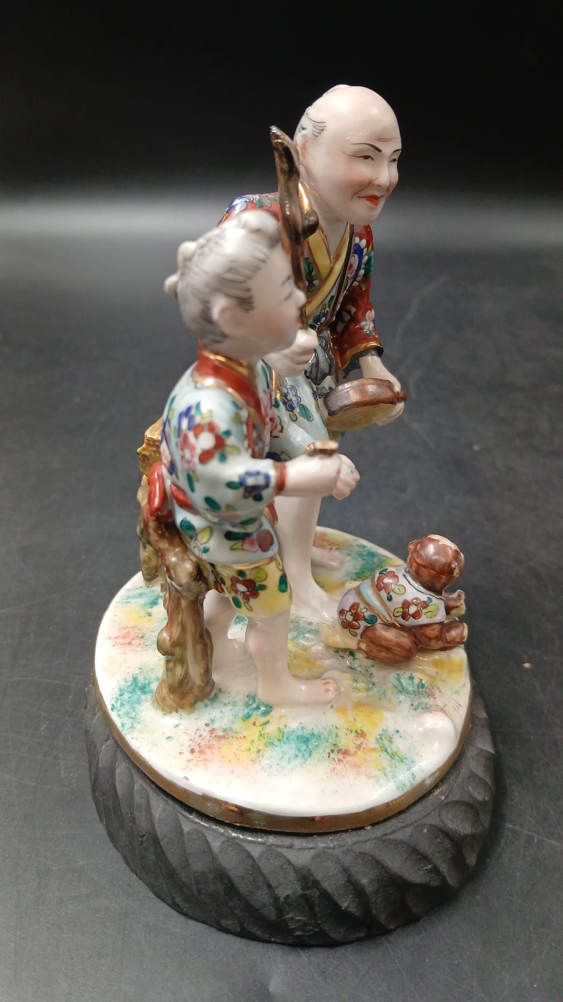Grupo escultórico em porcelana com duas figuras asiáticas e um pequeno animal, sobre base de madeira